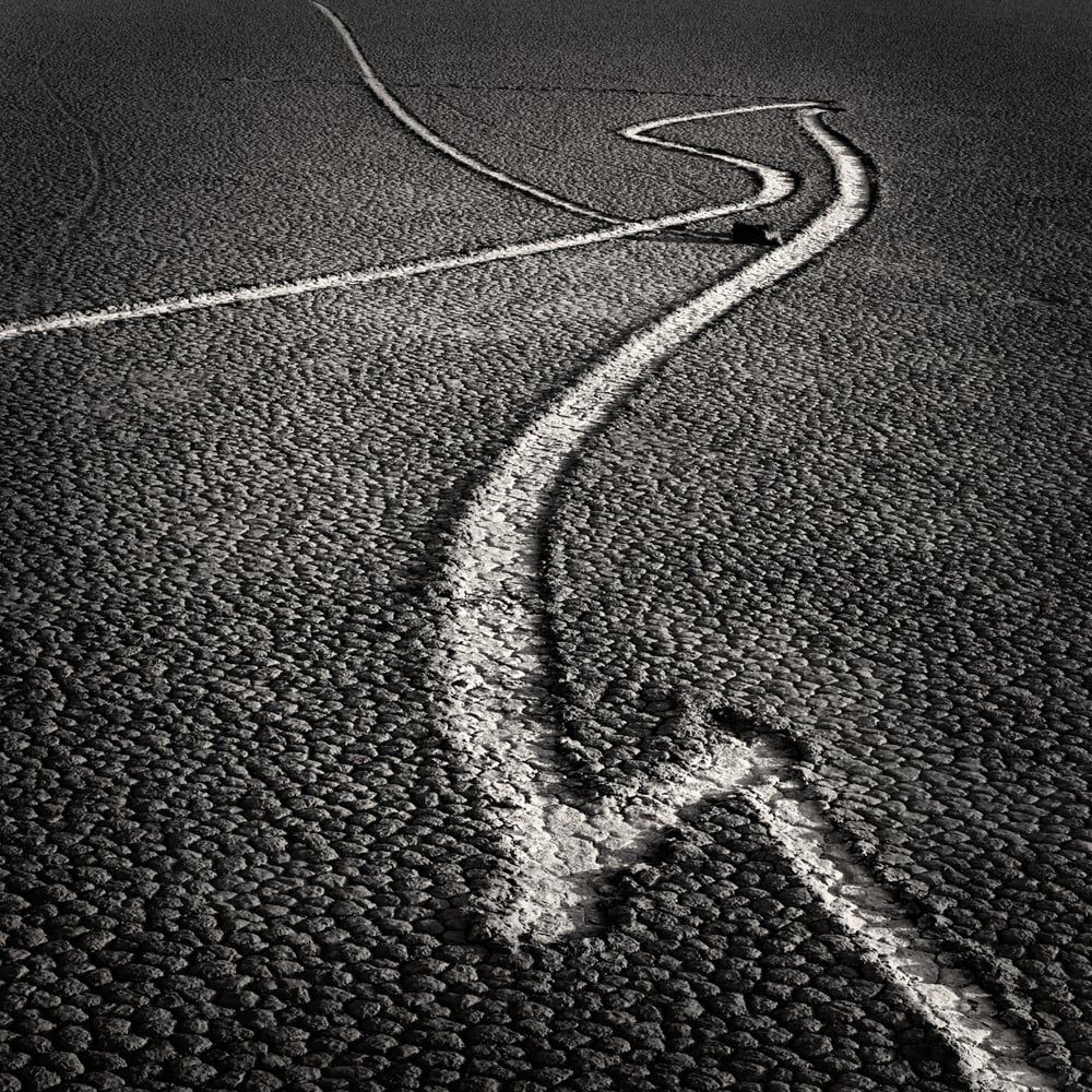 A sailing stone and its winding track across the cracked mud of a desert playa, black and white, by Michael E. Gordon
