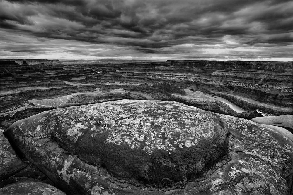 A lichen-covered boulder above a vast canyon vista under a dramatic stormy sky, black and white, by Bruce Hucko