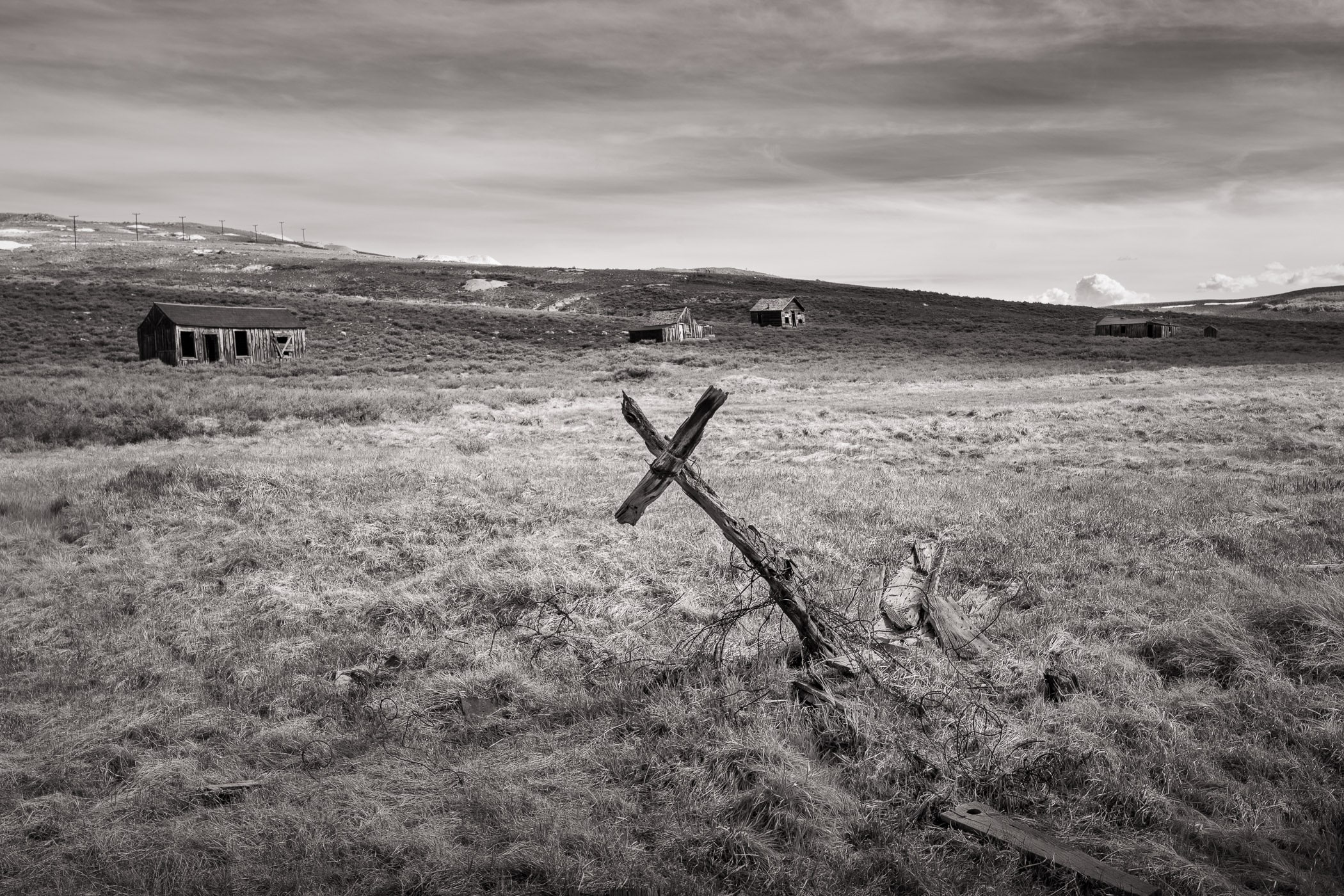 A leaning fence cross and weathered buildings on the prairie at Bodie, black and white, by Chuck Kimmerle